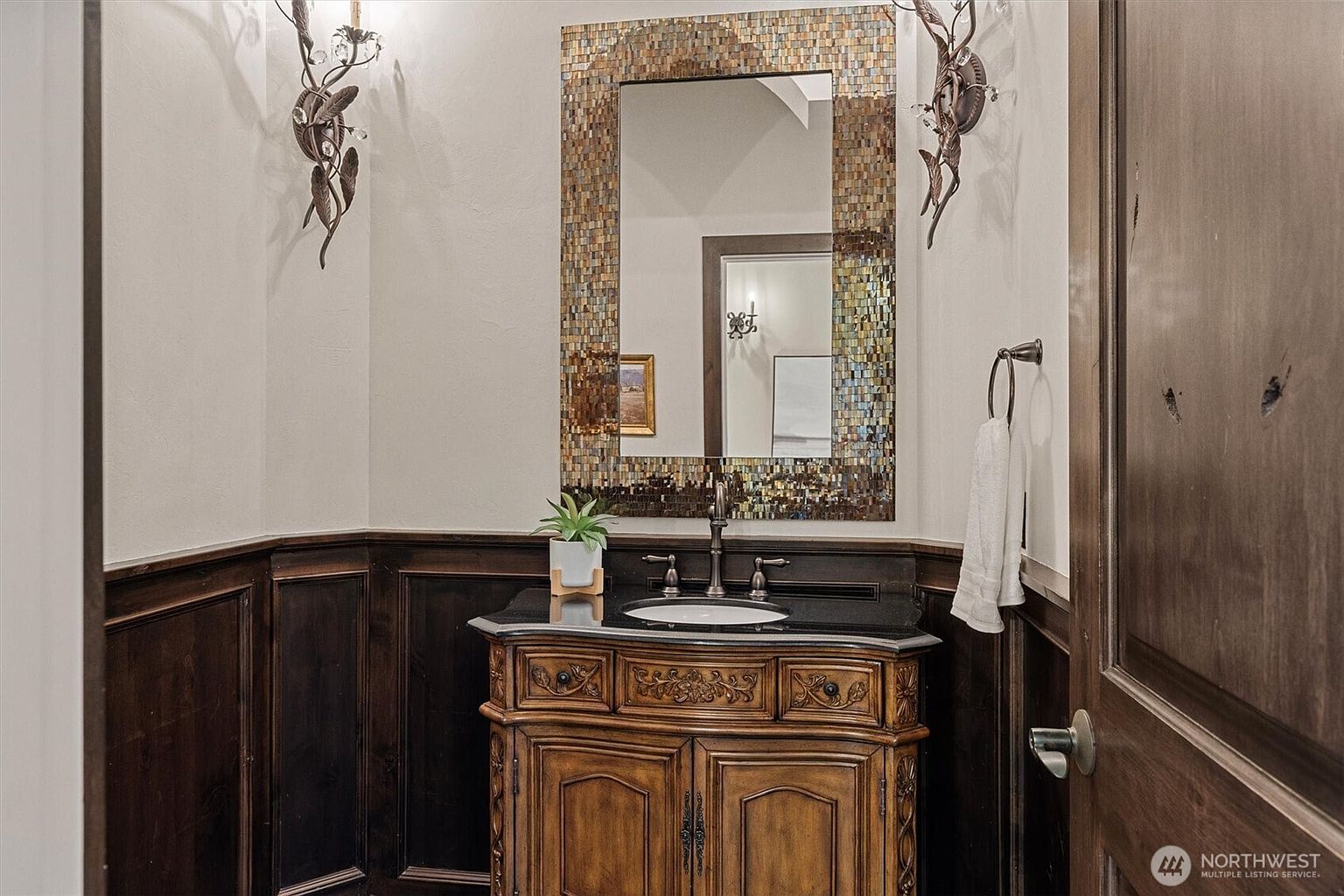 This is a well-appointed guest bathroom featuring dark wood paneling and a unique vanity with a black countertop. A mosaic-framed mirror hangs above the sink, flanked by decorative sconces. The room exudes a sense of classic elegance and attention to detail.