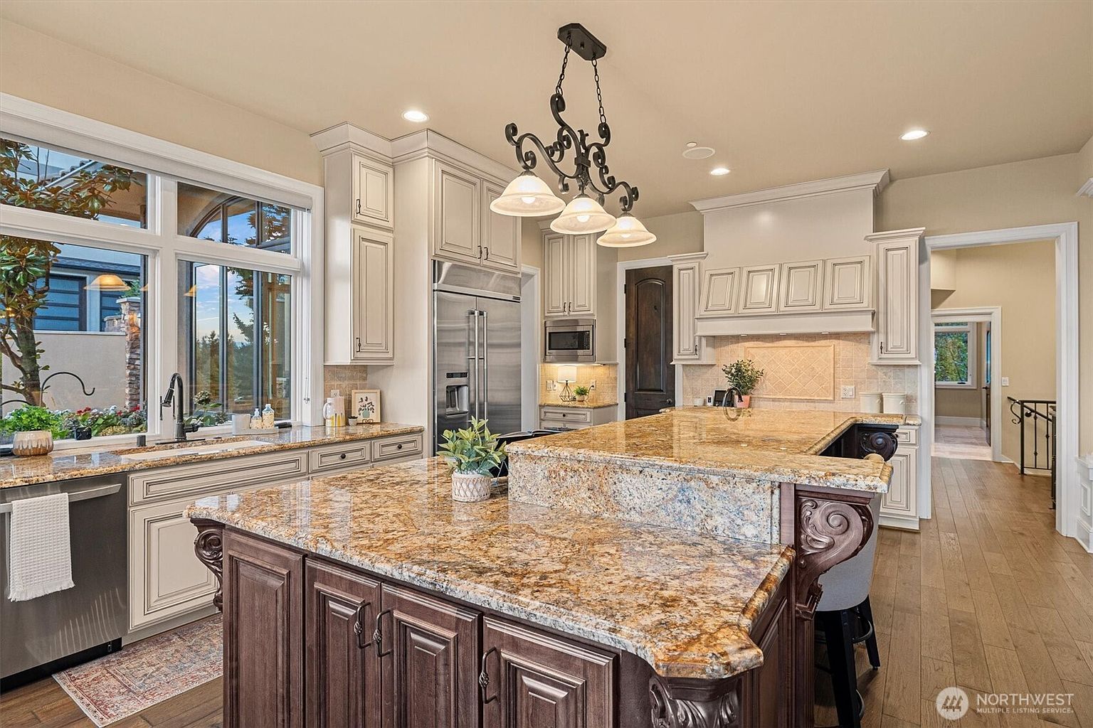 This is a well-lit kitchen featuring a large island with a granite countertop and dark wood cabinetry. The kitchen also includes white cabinets, stainless steel appliances, and a window overlooking the exterior. A decorative chandelier hangs above the island, adding a touch of elegance to the space.
