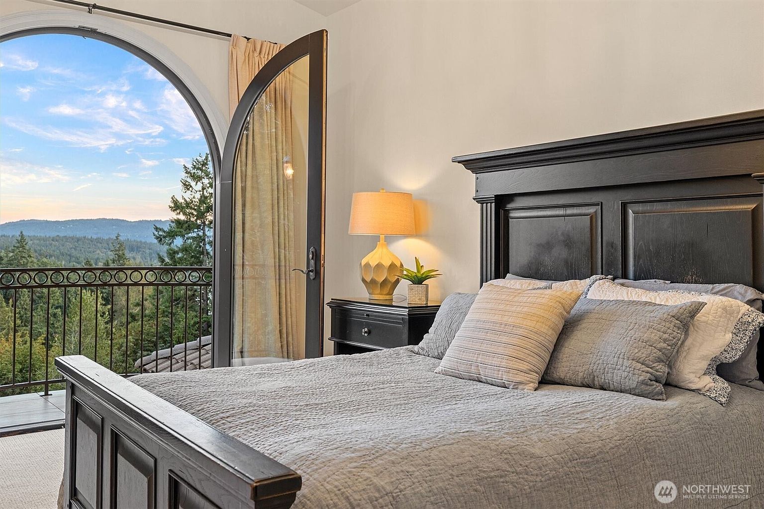 This is a primary bedroom featuring a dark wood bed frame with a gray quilted bedspread and several pillows. A matching nightstand holds a lamp and a small plant. An arched doorway leads to a balcony with a scenic view of trees and mountains, creating a luxurious and serene atmosphere.