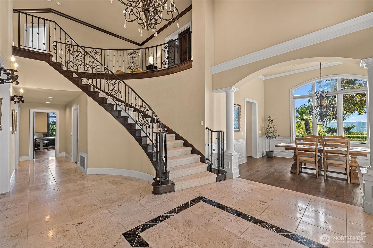 This grand foyer features a sweeping staircase with wrought iron railings, a chandelier, and travertine flooring with a decorative border. The space is open and airy, leading to a dining area with a large window offering natural light and views. The overall impression is one of luxury and elegance.