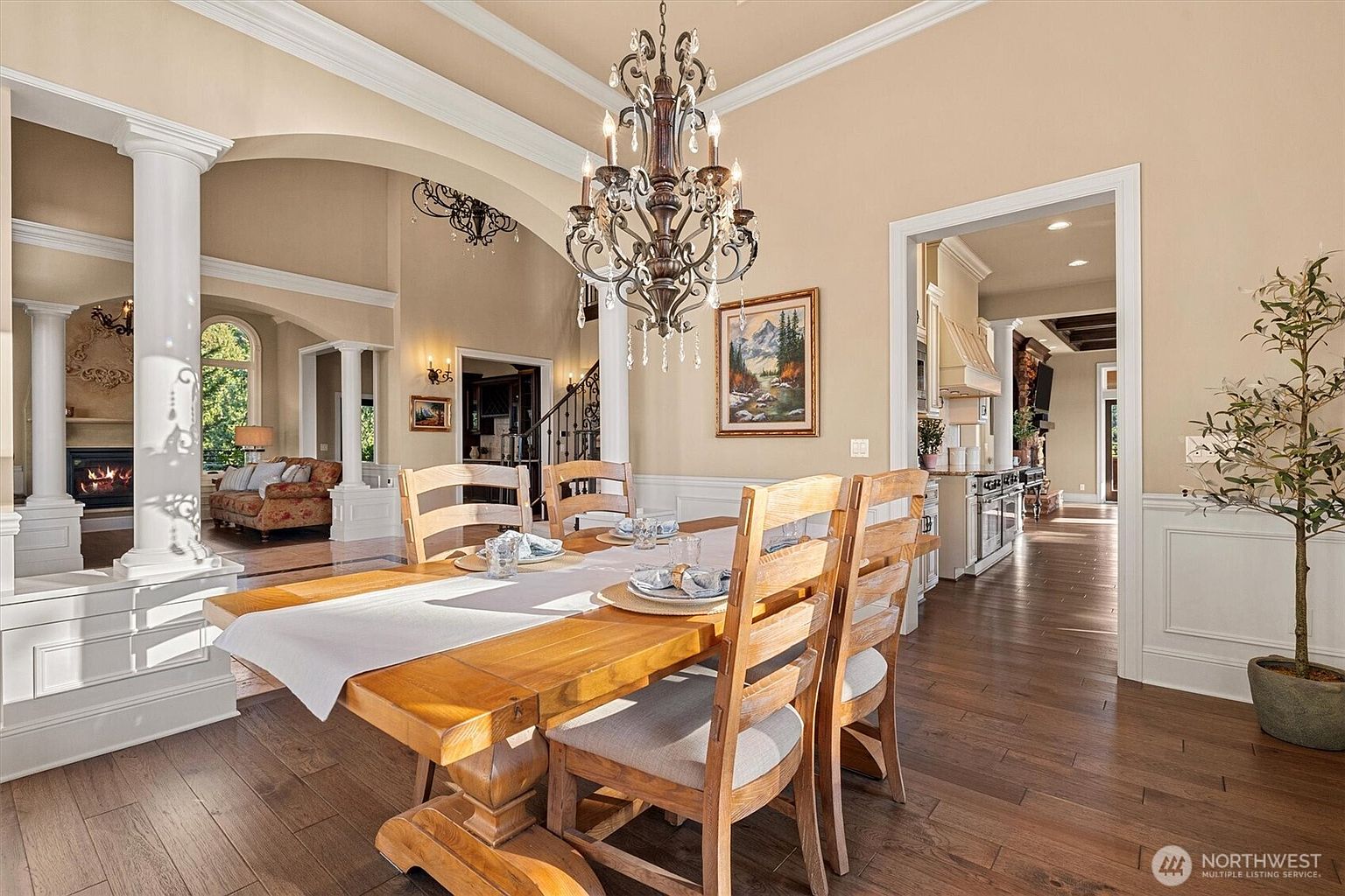 This is an interior shot of a dining room featuring a large wooden table set for a meal, illuminated by an ornate chandelier. The room is decorated in neutral tones with white trim and hardwood floors, creating a warm and inviting atmosphere. The open layout provides views into the living room and kitchen, enhancing the sense of spaciousness.