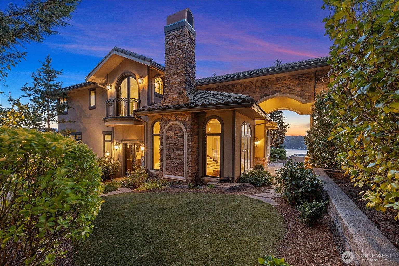 This is a stunning front view of a luxurious home at dusk, showcasing its architectural details and landscaping. The stone facade, arched windows, and prominent chimney create a sense of grandeur and elegance. The well-manicured lawn and surrounding greenery add to the property's curb appeal, making it an inviting and impressive residence.
