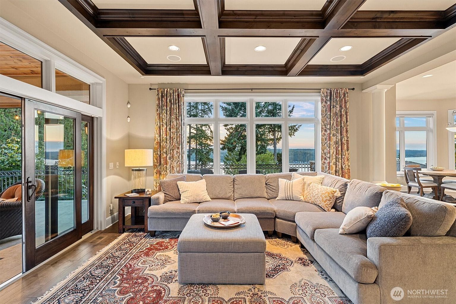This is an interior shot of a luxurious living room featuring a large, comfortable sectional sofa with an ottoman, positioned on an ornate area rug. The room is well-lit by natural light streaming through a large window with a view of the water and trees, complemented by stylish curtains. The coffered ceiling adds an elegant touch, and the overall impression is one of comfort and sophistication.