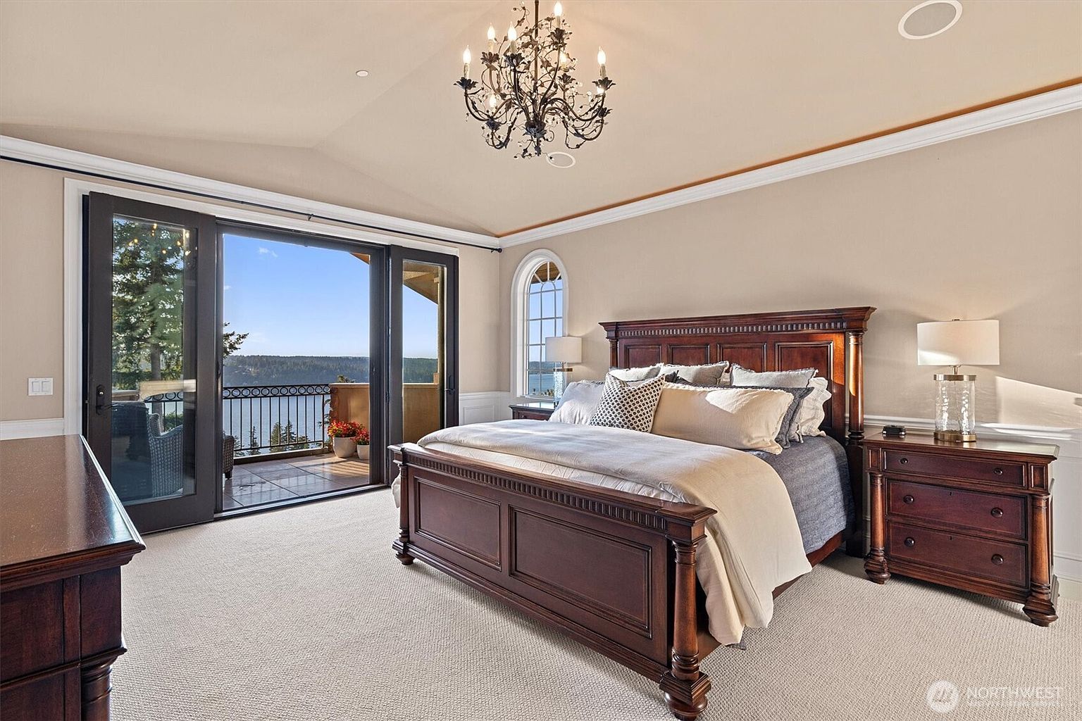 This is a luxurious primary bedroom featuring a dark wood, four-poster bed with plush bedding and decorative pillows. The room has a neutral color palette with elegant crown molding and a chandelier. Sliding glass doors lead to a balcony with a scenic water view, enhancing the room's appeal and creating a serene atmosphere.
