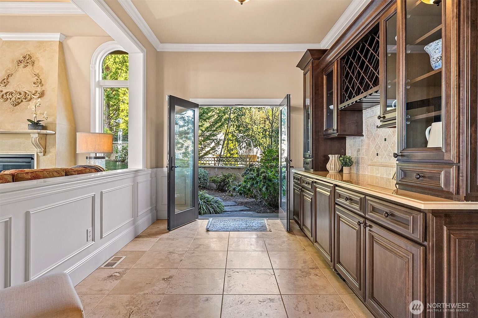 This interior shot showcases a hallway with tiled flooring leading to an open doorway that reveals a lush garden. On the right, dark wood cabinetry with glass doors adds a touch of elegance, while the left side features white wainscoting and a glimpse into another room. The overall impression is one of refined taste and seamless indoor-outdoor living.