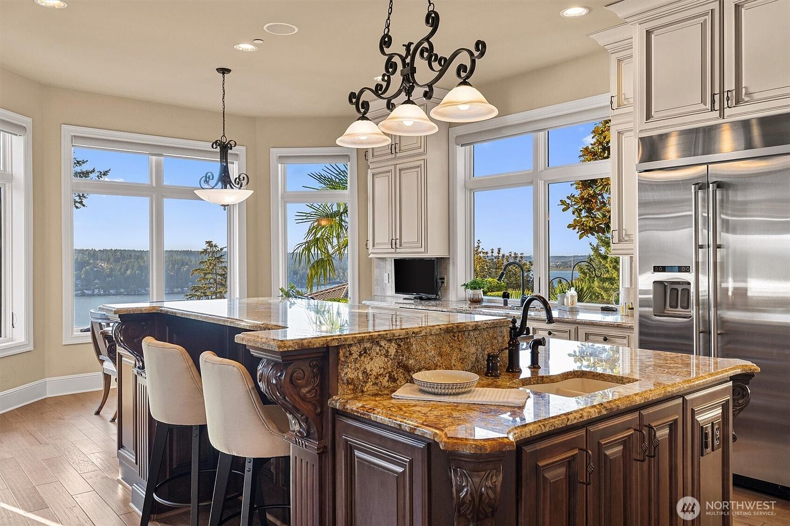 This is a well-lit kitchen featuring a large island with granite countertops and dark wood cabinetry. The kitchen has stainless steel appliances and large windows that offer a scenic view of the water. Ornate light fixtures hang above the island, adding a touch of elegance to the space.