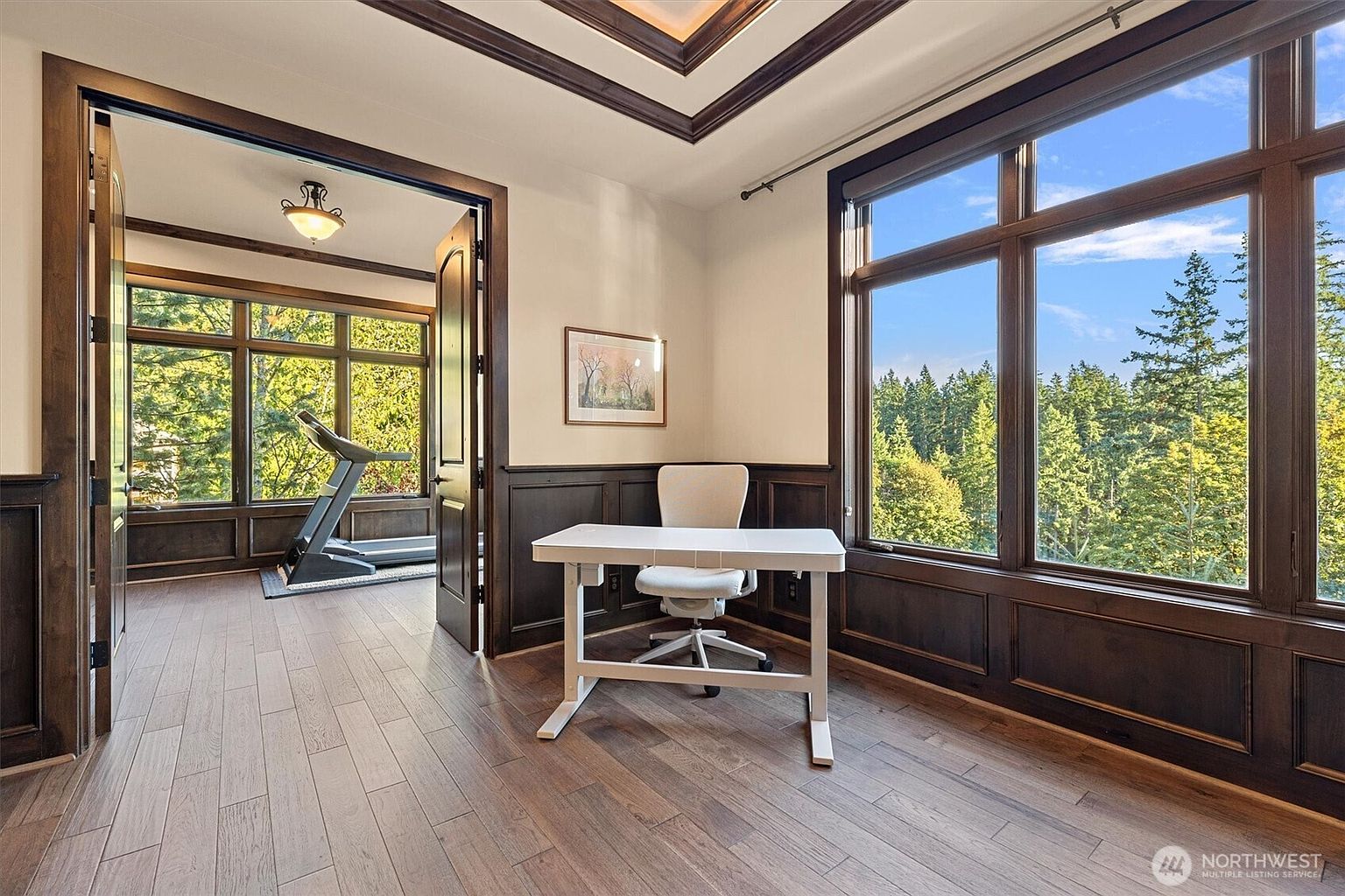 This is an interior shot of a home office featuring hardwood floors, dark wood trim, and a large window offering a view of the trees outside. A white desk and chair are positioned in the center of the room, and an open doorway leads to another room with a treadmill. The room exudes a sense of sophistication and tranquility, ideal for a productive workspace.