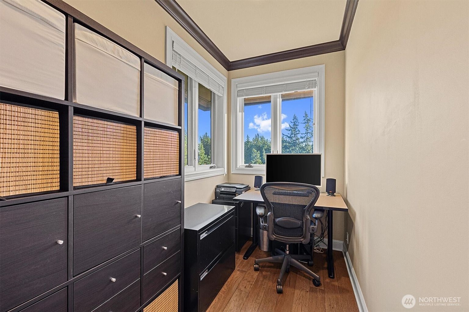 This is an interior shot of a home office. The room features a desk with a computer, an ergonomic chair, and storage units. Natural light streams in through the windows, creating a bright and inviting workspace.