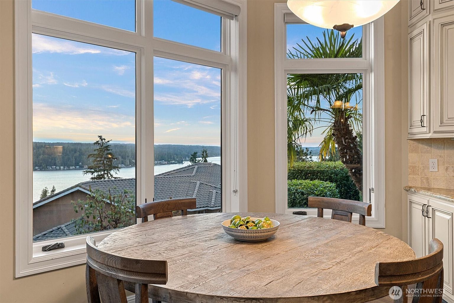 This interior shot showcases a dining area with a large, round wooden table as the focal point, complemented by wooden chairs. Natural light floods the room through expansive windows, offering scenic views of the water and lush greenery. The overall ambiance is bright, inviting, and highlights the property's desirable location and architectural design.