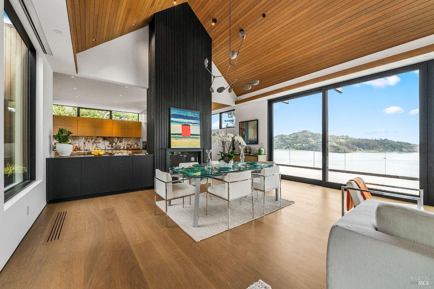 This open-concept dining area features a glass-topped table surrounded by modern chairs, set against a striking black vertical-slat chimney breast that anchors the space. The room boasts a vaulted wood-paneled ceiling and expansive glass walls that provide panoramic views of the water and hills beyond. A sleek, contemporary kitchen with warm-toned cabinetry is visible in the background, creating a cohesive and sophisticated living environment.