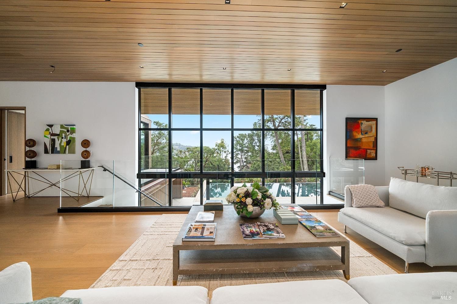 This sophisticated living room features a warm, wood-paneled ceiling and light-toned hardwood flooring, creating an inviting and airy atmosphere. A large, grid-patterned window wall serves as the focal point, framing a scenic view of the surrounding trees and a glimpse of a swimming pool below. The space is furnished with plush white sofas and a substantial, low-profile coffee table, complemented by minimalist art and a glass-railed staircase that adds to the modern, open-concept design.