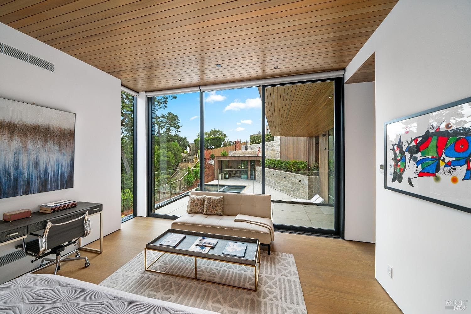 This modern, light-filled office space features a sleek desk setup and a comfortable seating area arranged on a patterned area rug. The room is defined by expansive floor-to-ceiling glass walls that provide a seamless transition to the outdoor patio and pool area. A warm wood-paneled ceiling adds a natural, sophisticated touch to the minimalist interior design.