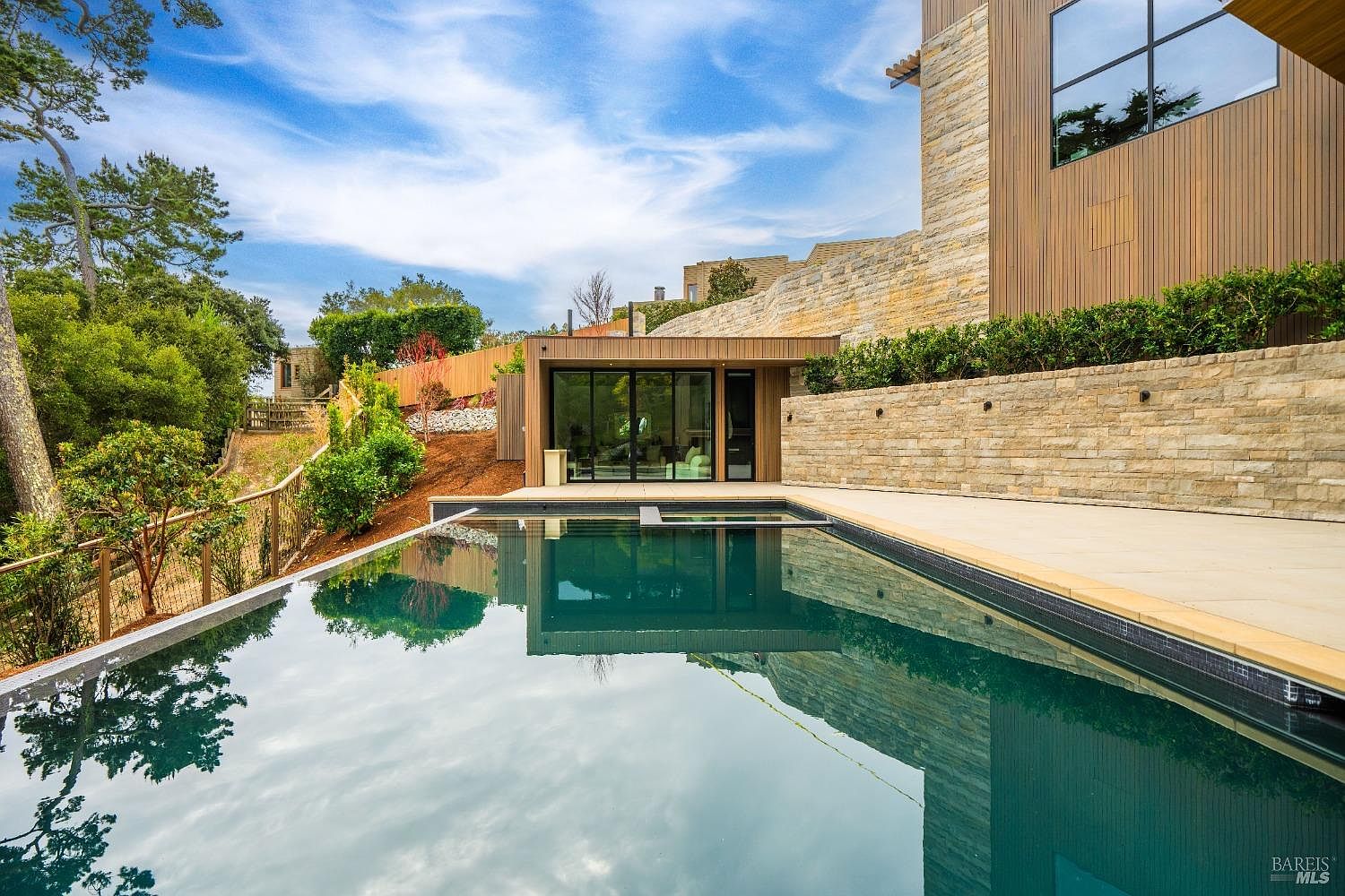 This modern outdoor space features a sleek, dark-tiled swimming pool that reflects the surrounding architecture and blue sky. A stone-clad retaining wall creates a tiered effect, leading up to a contemporary structure with large glass doors and wood-paneled siding. The scene is set against a backdrop of mature trees and a hillside, offering a serene and luxurious atmosphere.