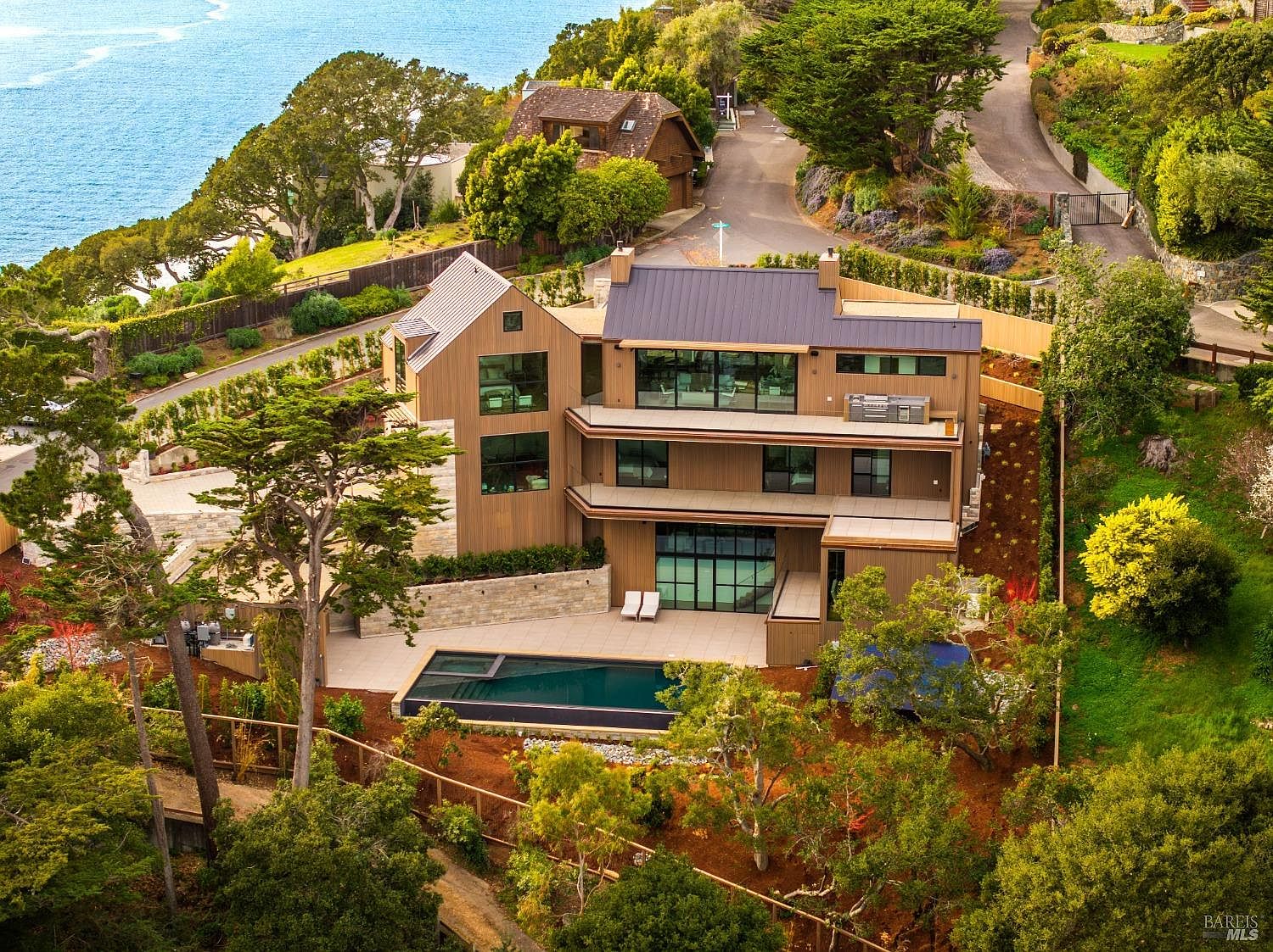 This aerial perspective captures a modern, multi-level residence nestled on a hillside overlooking the ocean. The home features vertical wood siding, expansive glass windows, and multiple tiered balconies that provide seamless indoor-outdoor living. A sleek, dark-bottomed swimming pool sits on the lower patio, surrounded by lush landscaping and mature trees, creating a serene and luxurious coastal retreat.