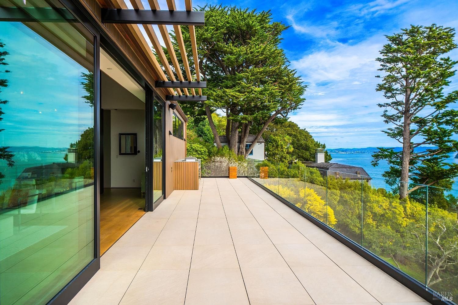 This expansive, modern deck features large-format light-toned tiles and a seamless glass railing that provides an unobstructed view of the surrounding trees and the distant bay. A wide sliding glass door connects the indoor living space to the outdoor area, which is partially shaded by a slatted wood pergola. The setting offers a serene, elevated perspective, emphasizing the home's integration with the natural landscape.