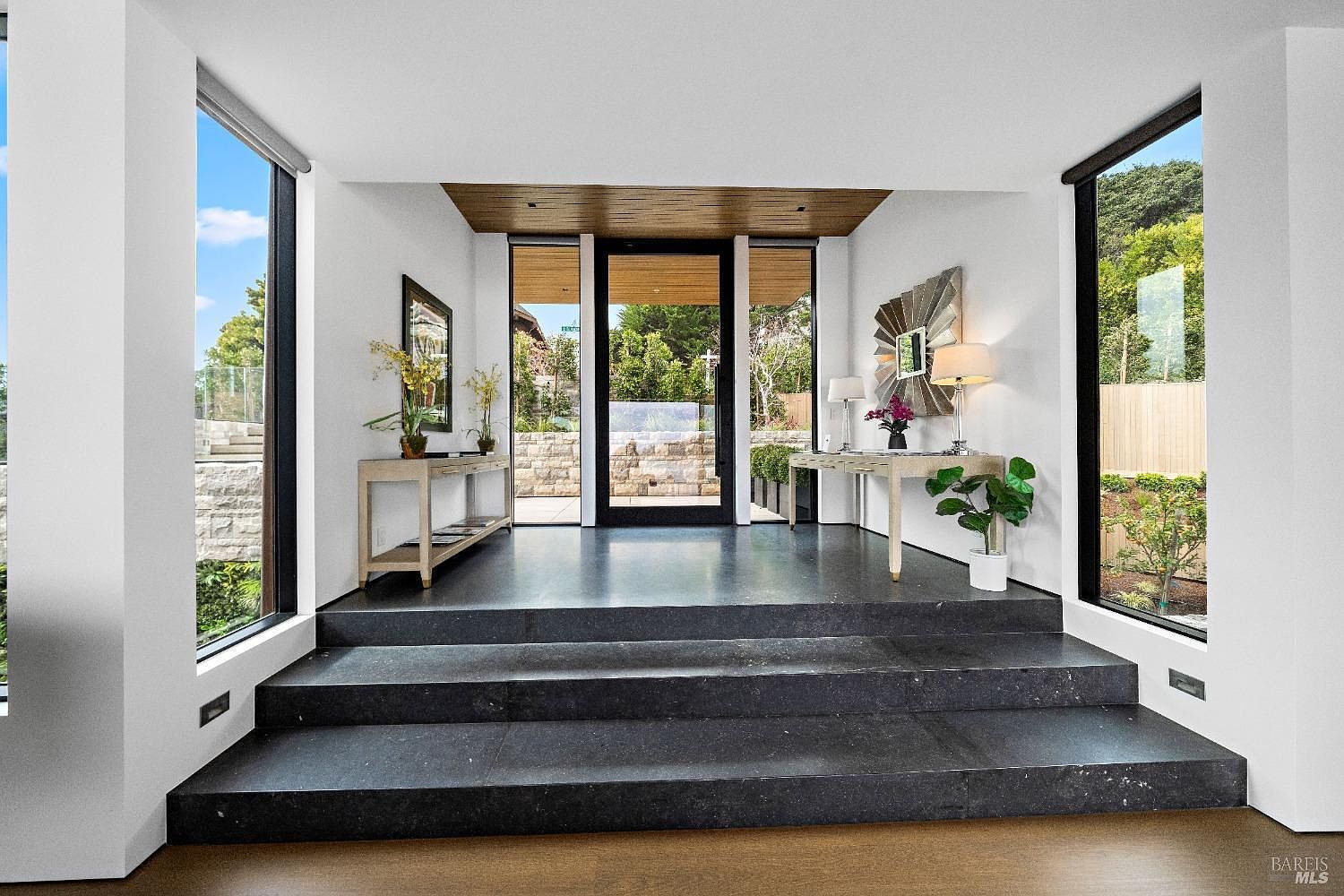 This sophisticated entryway features a set of wide, dark stone steps leading up to a glass-paneled front door framed by large windows that offer a clear view of the outdoor landscape. The space is symmetrically designed with light-toned console tables on either side, topped with decorative mirrors, lamps, and floral arrangements. The clean white walls and warm wood-paneled ceiling create a bright, modern, and welcoming atmosphere.