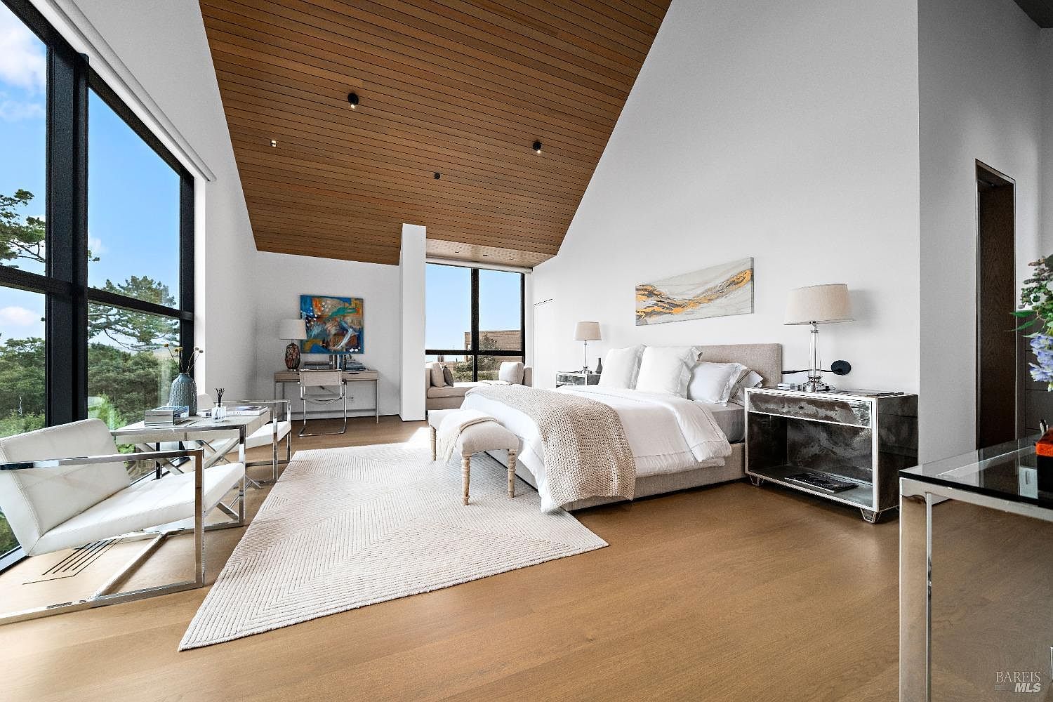 This spacious primary bedroom features a dramatic vaulted ceiling with warm wood paneling and large windows that provide expansive views of the surrounding landscape. The room is anchored by a plush bed with neutral bedding, complemented by mirrored nightstands and a sleek desk area. A light-colored area rug sits atop the light wood flooring, creating a bright, airy, and sophisticated atmosphere.