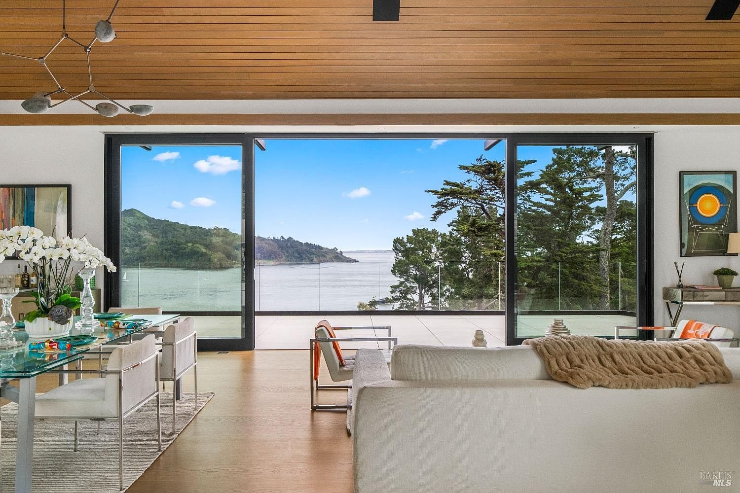 This open-concept living and dining area features a sophisticated, modern aesthetic with light wood flooring and a warm wood-paneled ceiling. The space is defined by expansive floor-to-ceiling glass sliding doors that frame a panoramic view of the bay and surrounding hills. A plush white sofa and contemporary dining set anchor the room, while a unique sculptural chandelier adds a touch of artistic flair to the bright, airy interior.