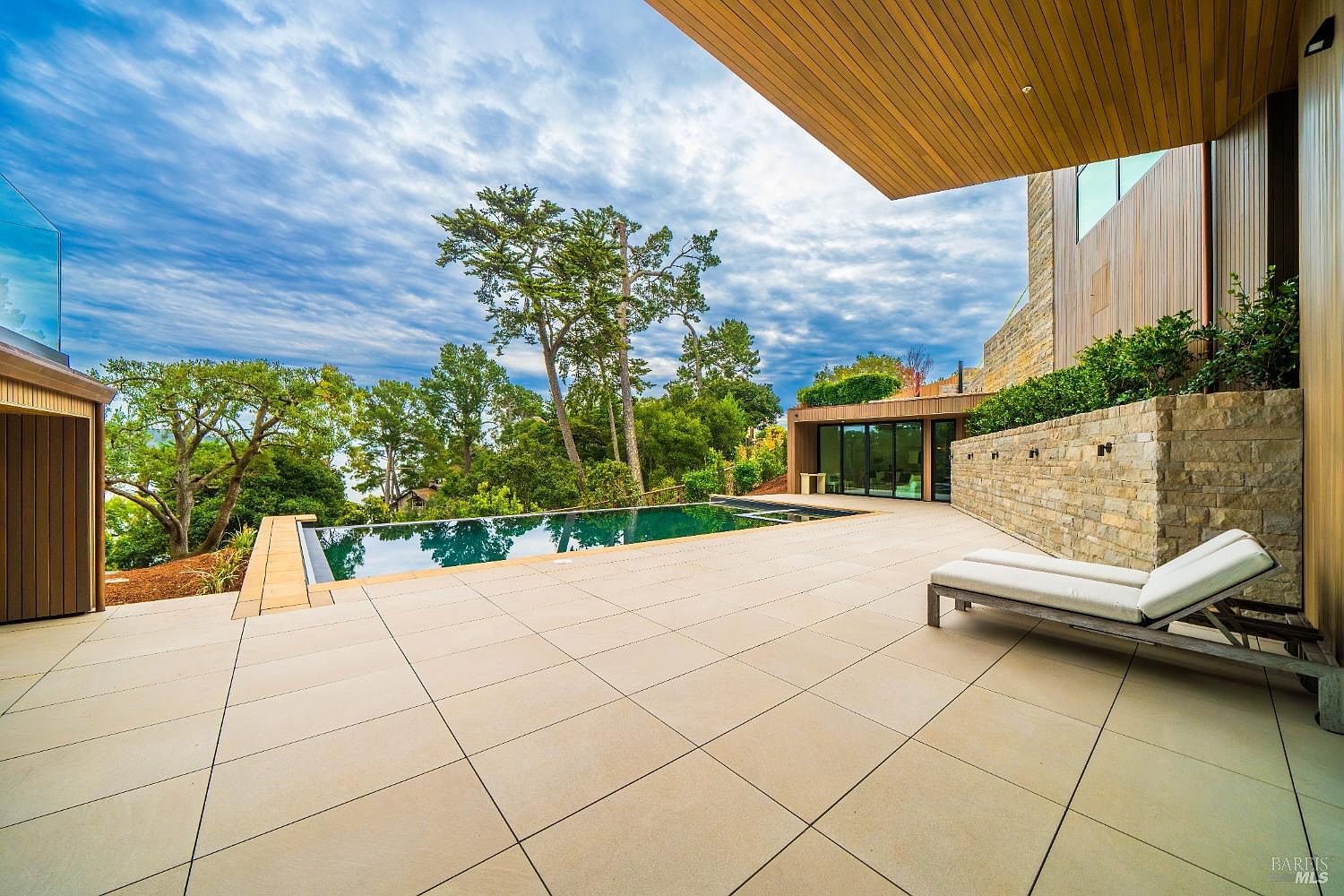 This luxurious outdoor living space features a sleek, rectangular infinity-edge pool that overlooks a lush, tree-lined landscape. A spacious stone-paved patio provides ample room for relaxation, highlighted by a modern lounge chair positioned under the architectural overhang of the home. The scene is framed by a textured stone wall and large glass doors, creating a seamless transition between the contemporary indoor and outdoor environments.