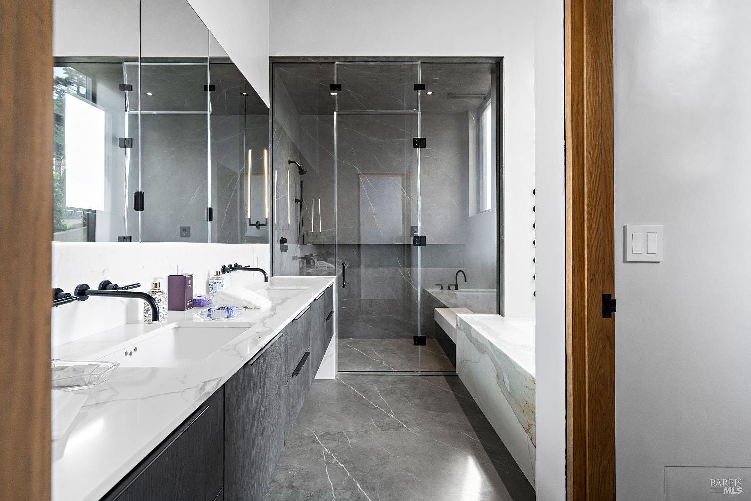 This modern primary bathroom features a sleek, dark-toned vanity with a light-colored stone countertop and dual wall-mounted faucets. A glass-enclosed shower with large-format stone tiles sits adjacent to a deep soaking tub, creating a clean and sophisticated aesthetic. The perspective looks into the bathroom from a doorway, highlighting the seamless integration of high-end fixtures and reflective surfaces.
