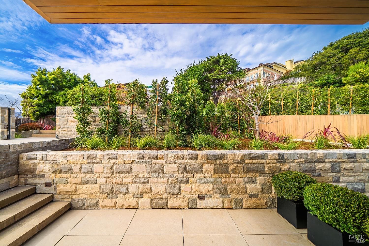 This outdoor space features a clean, modern patio paved with large light-toned tiles, leading up to a tiered stone retaining wall. The garden area above is landscaped with young trees, ornamental grasses, and shrubs, framed by a wooden fence and a stone wall. The perspective is from a covered area looking out, emphasizing the structured, contemporary design of the terraced landscape.