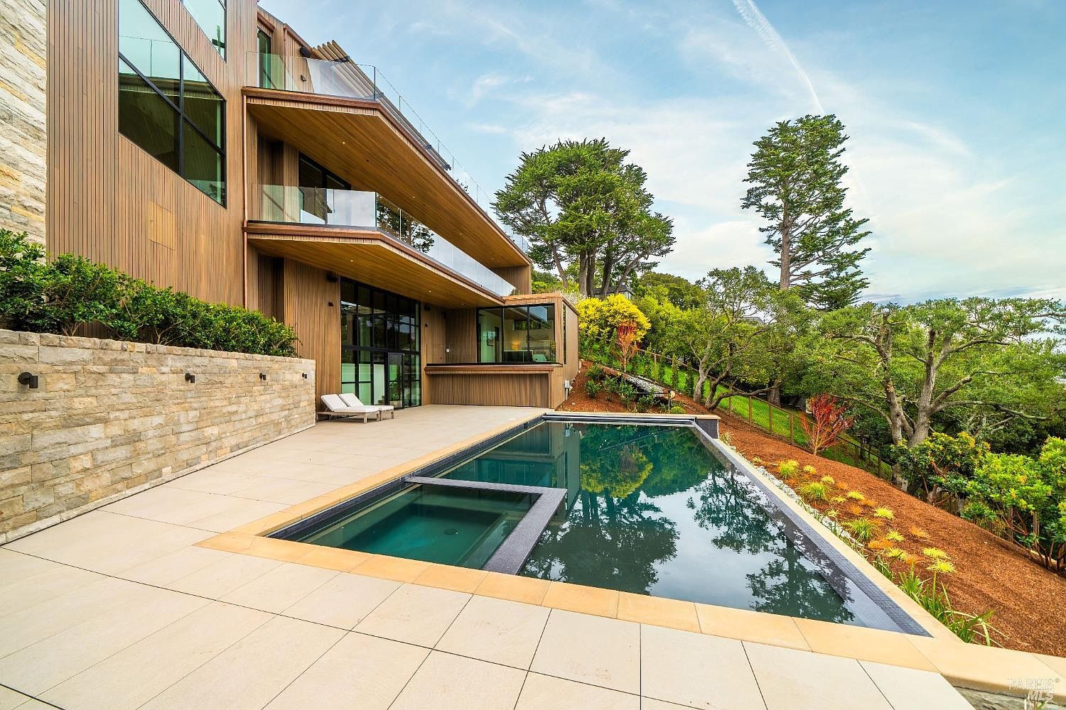 This modern luxury residence features a multi-level exterior design with warm-toned wood siding and expansive glass walls. A sleek, rectangular infinity-edge pool with an integrated spa sits on a spacious stone-paved patio, offering a seamless transition to the surrounding hillside landscape. The scene captures a sophisticated outdoor living space that emphasizes clean lines, natural textures, and a serene connection to the outdoors.
