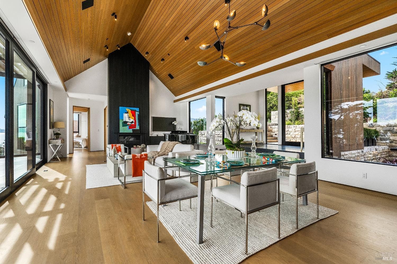 This open-concept living and dining area features a dramatic vaulted ceiling with warm wood paneling and a modern, multi-armed chandelier. A glass-topped dining table sits in the foreground, while a living area with a tall, dark-paneled chimney breast and contemporary furniture occupies the background. Large windows and sliding glass doors provide abundant natural light and views of the outdoor landscape, creating a bright and airy atmosphere.