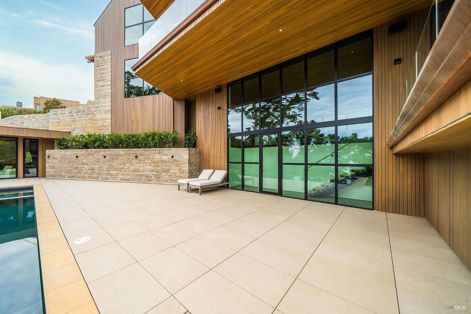 This modern outdoor living space features a spacious stone-tiled patio adjacent to a swimming pool. The area is defined by a striking architectural overhang clad in warm-toned wood, which shelters a wall of large glass panels that provide a seamless transition to the interior. A low stone retaining wall with integrated lighting adds texture and depth, while two lounge chairs offer a comfortable spot to enjoy the serene, contemporary setting.
