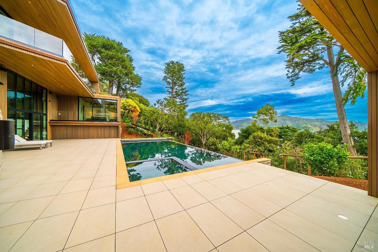 This modern architectural home features a sprawling, light-toned stone patio that seamlessly integrates with an infinity-edge pool overlooking a lush, forested landscape and distant hills. The structure utilizes extensive glass walls and warm wood siding to create a sophisticated indoor-outdoor living experience. The perspective captures the expansive deck area, emphasizing the tranquil connection between the luxury residence and the natural environment.