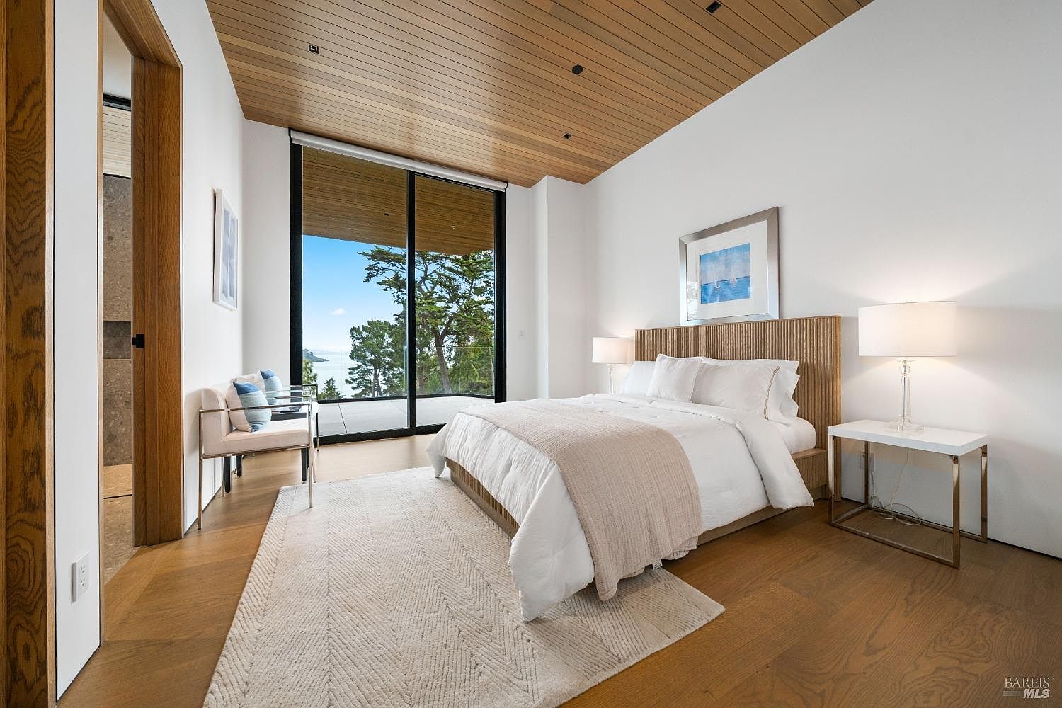 This serene primary bedroom features a warm wood-paneled ceiling and light wood flooring, creating a cohesive and natural aesthetic. A large bed with a textured headboard serves as the focal point, complemented by a neutral color palette and a soft area rug. The room is anchored by floor-to-ceiling glass sliding doors that provide an expansive view of the outdoors and lead to a private balcony.