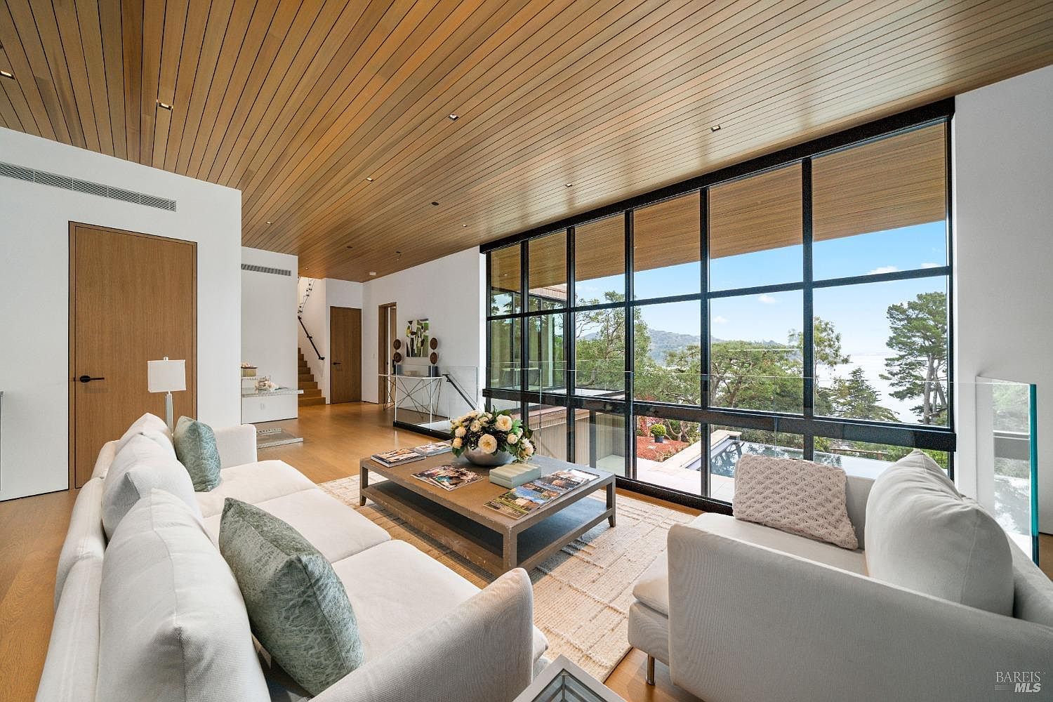 This sophisticated living room features a warm wood-paneled ceiling and light wood flooring, creating a cohesive and inviting atmosphere. A large, grid-patterned window wall provides an expansive view of the lush outdoor landscape and distant water. The space is furnished with a plush white sofa and a central coffee table, emphasizing a clean, modern aesthetic with a focus on natural light and indoor-outdoor connectivity.