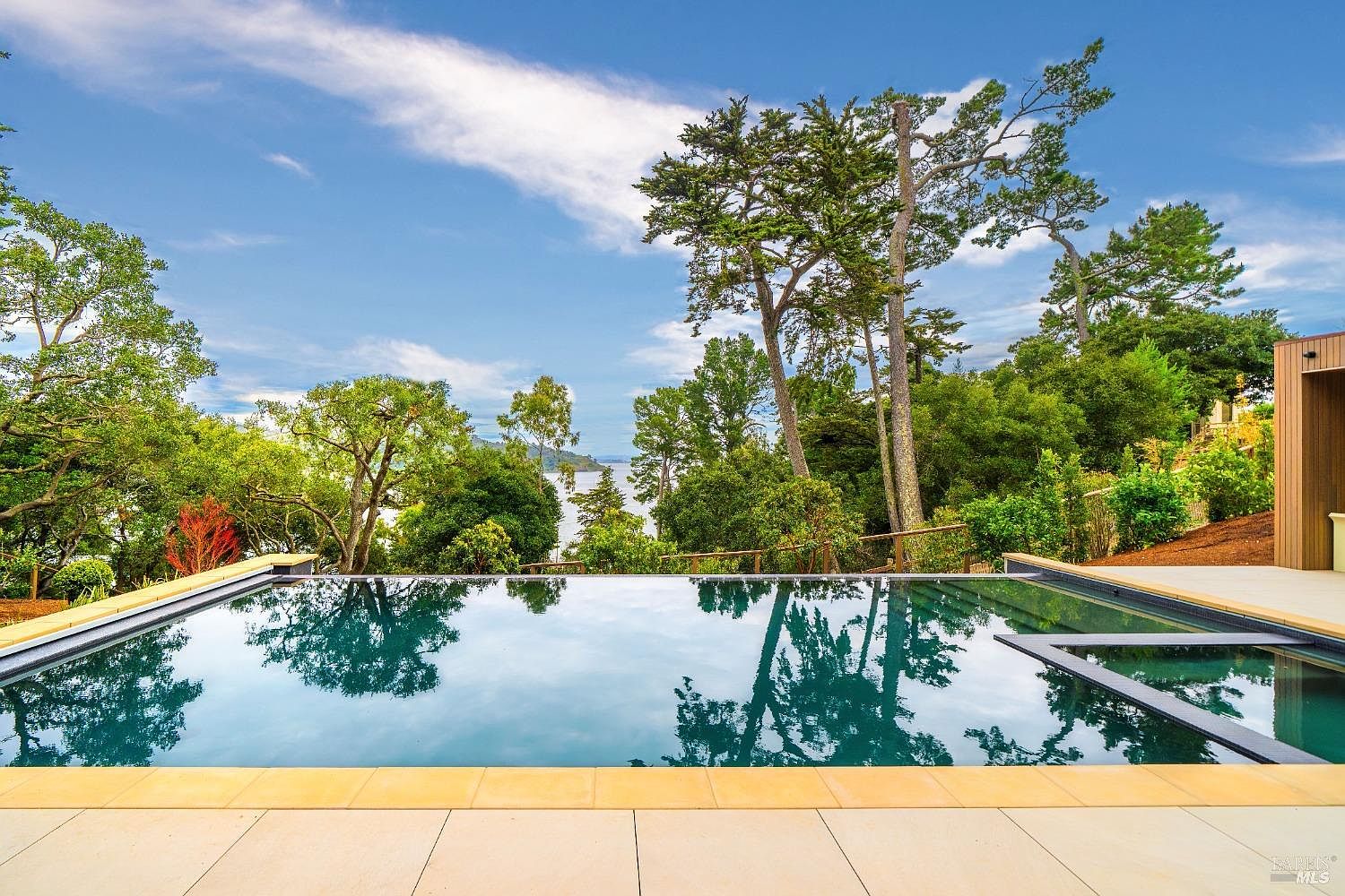 This image captures a serene infinity-edge swimming pool overlooking a lush, tree-lined landscape and a distant body of water. The pool deck is paved with light-colored stone tiles, providing a clean, modern foreground that contrasts with the natural greenery and blue sky. The perspective is from the edge of the pool, emphasizing the seamless transition between the water and the scenic horizon.