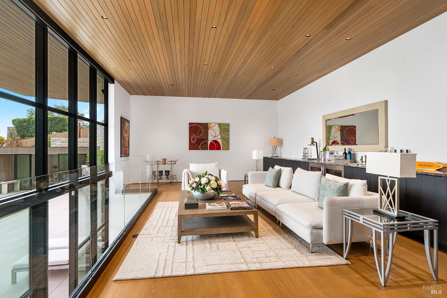 This sophisticated living room features a warm wood-paneled ceiling with recessed lighting and light wood flooring. A large white sectional sofa and a central coffee table create a comfortable seating area, complemented by a glass-railed balcony overlooking an outdoor space. The room is accented by a wet bar area with dark cabinetry and a large mirror, providing a sleek and modern aesthetic.