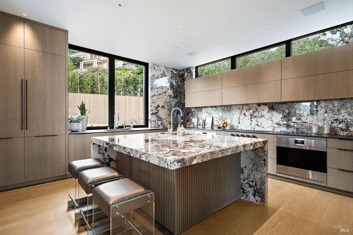 This modern kitchen features a large central island with a waterfall-edge stone countertop and a textured base, complemented by three sleek stools. The space is defined by light wood cabinetry, a striking patterned stone backsplash, and expansive windows that provide a bright, airy atmosphere. The overall aesthetic is sophisticated and contemporary, showcasing high-end finishes and a seamless, open-concept design.