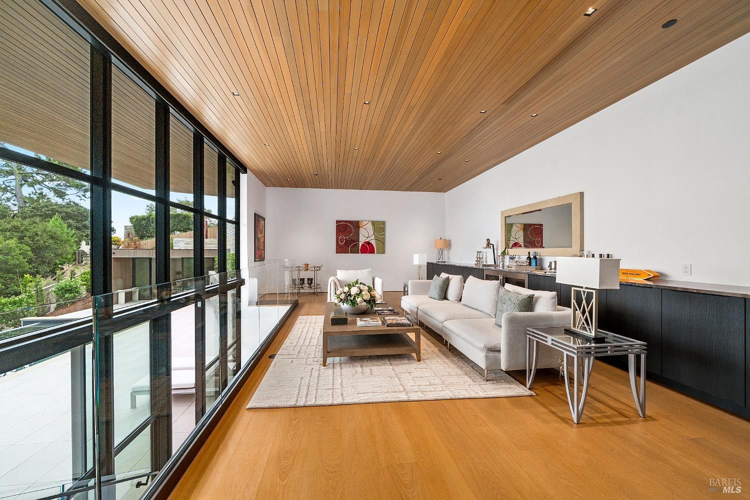 This modern living room features a striking wood-paneled ceiling and expansive floor-to-ceiling windows that provide a seamless connection to the outdoor space. The room is furnished with a neutral-toned sofa, a central coffee table, and a geometric-patterned area rug, all set against a backdrop of clean white walls. A sleek, dark-toned console runs along the wall, complemented by a decorative mirror and contemporary lighting fixtures, creating a sophisticated and airy atmosphere.