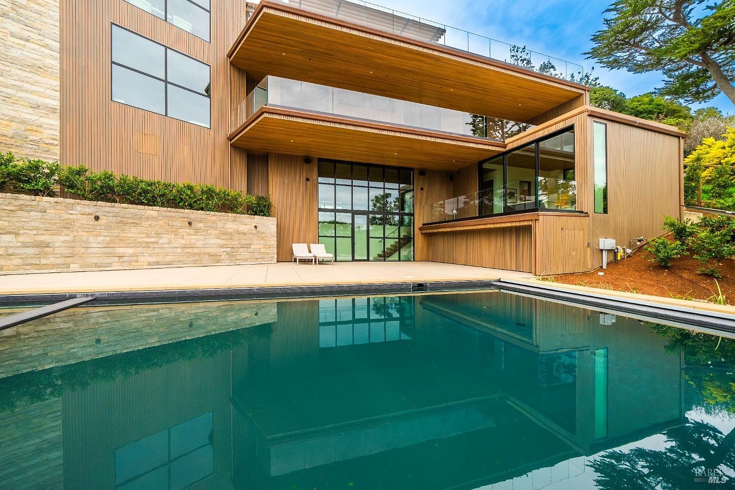 This modern architectural residence features a striking combination of vertical wood siding and stone masonry, centered around a large, dark-bottom swimming pool. The home showcases expansive glass walls and a cantilevered upper balcony with a glass railing, creating a seamless indoor-outdoor aesthetic. A stone retaining wall lines the patio area, which is furnished with two lounge chairs, all set against a backdrop of mature trees and lush landscaping.