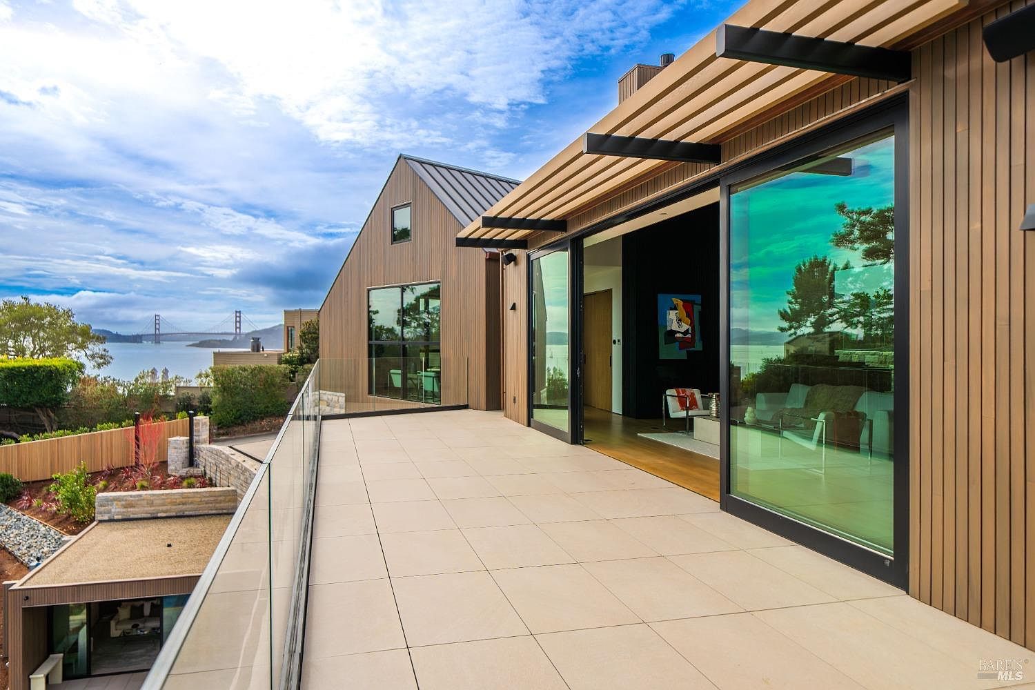 This expansive, light-toned tiled balcony offers a seamless indoor-outdoor transition through large sliding glass doors that open into a contemporary living space. The deck features a sleek glass railing that provides an unobstructed view of the bay and the Golden Gate Bridge in the distance. The architectural design incorporates warm-toned vertical wood siding and a modern pergola structure, creating a sophisticated and airy atmosphere.