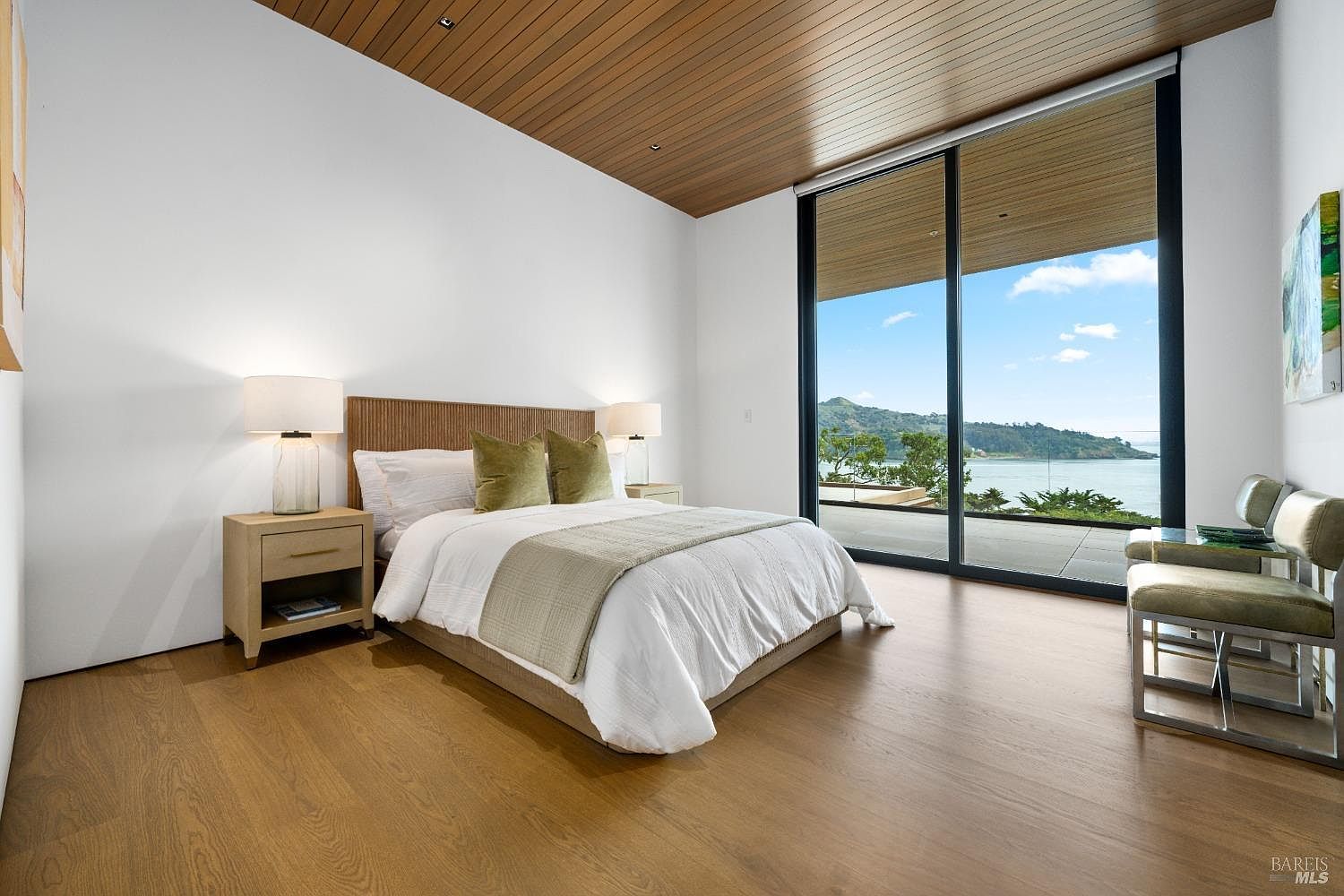 This modern primary bedroom features light wood flooring and a warm wood-paneled ceiling, creating a serene and sophisticated atmosphere. A plush bed with neutral bedding and olive-toned accent pillows serves as the focal point, flanked by two matching nightstands with contemporary lamps. Large floor-to-ceiling glass sliding doors provide an expansive, unobstructed view of the bay and distant hills, seamlessly blending indoor living with the natural landscape.