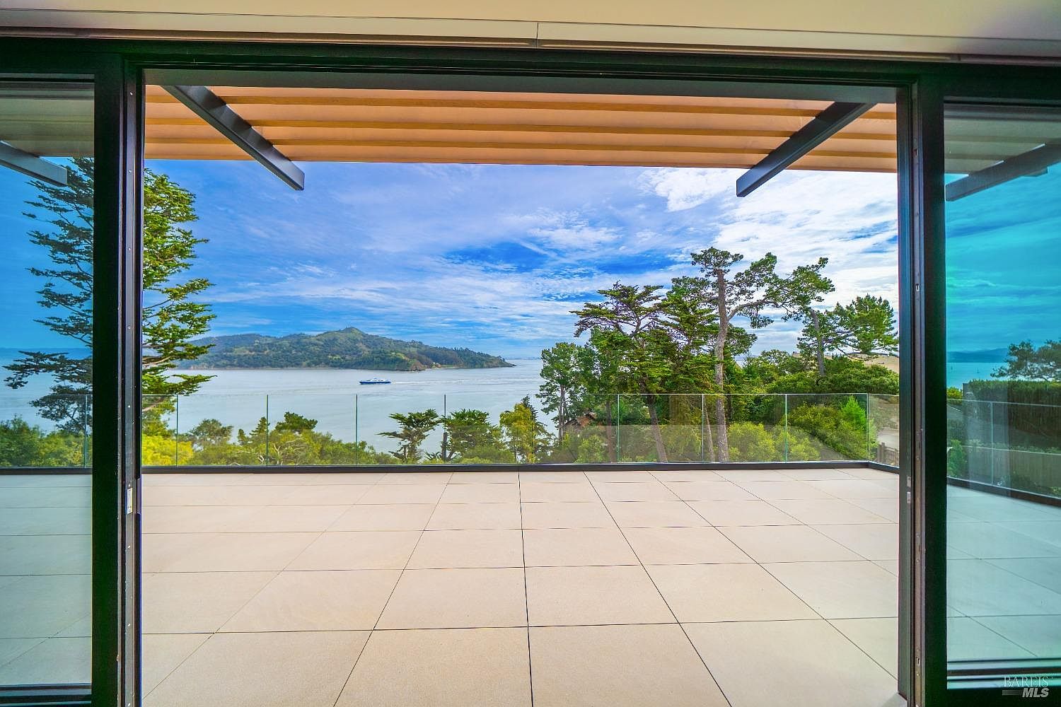 This image captures a spacious, modern balcony featuring large, light-colored square tiles and a sleek glass railing that provides an unobstructed view of the bay. The perspective is from inside looking out through wide-opening sliding glass doors, framing a stunning vista of the water, distant hills, and a passing boat under a bright, cloudy sky. The architectural design emphasizes a seamless transition between indoor and outdoor living spaces, highlighted by the clean lines of the overhead structural beams.