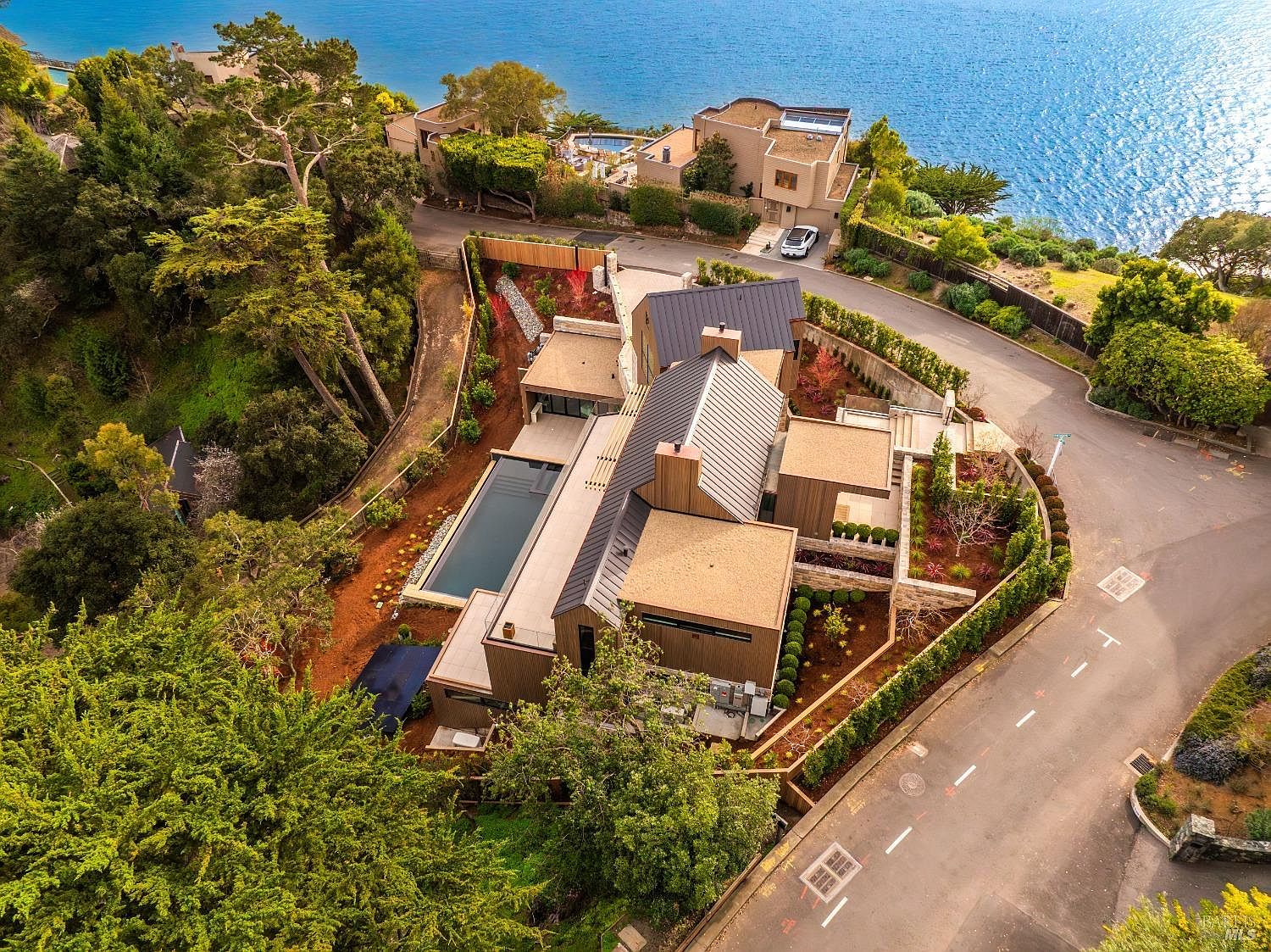This high-angle aerial view captures a modern, multi-level residence nestled on a coastal hillside overlooking the water. The home features a contemporary design with dark metal roofing, light-toned siding, and a rectangular swimming pool integrated into the landscaped grounds. The property is surrounded by lush trees and winding roads, emphasizing its secluded and scenic location.
