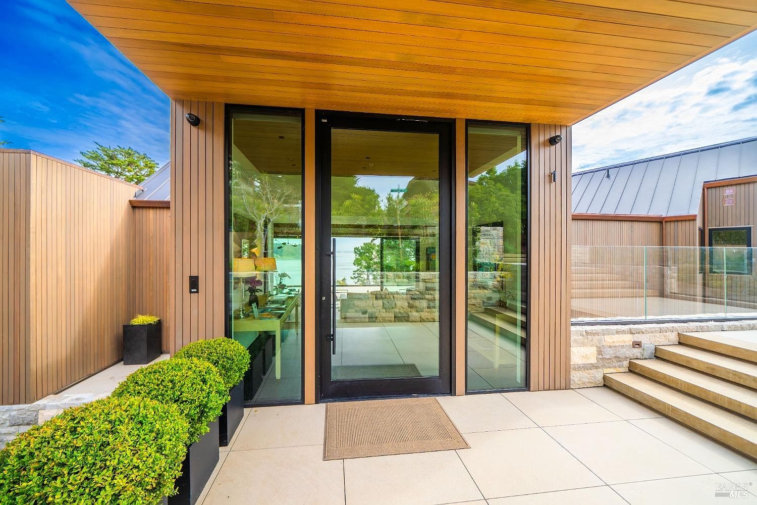This modern entryway features a prominent glass-paneled front door framed by large windows that offer a clear view through the home to the water beyond. The exterior is clad in vertical light wood siding, complemented by a wide, covered porch with large-format light-colored tiles and a set of stone steps leading to a glass-railed deck. The clean lines, minimalist aesthetic, and integration of natural materials create a sophisticated and welcoming first impression.