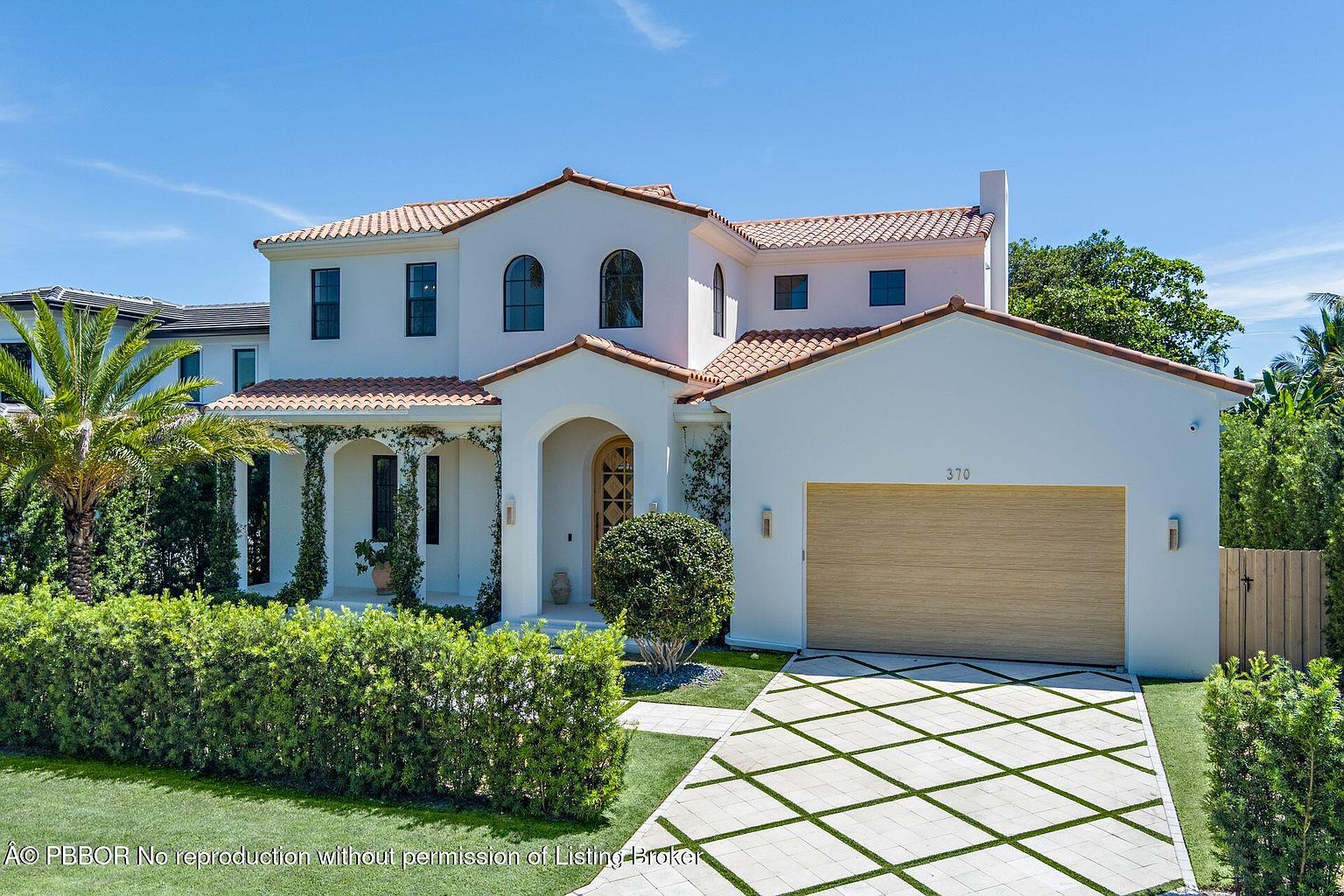 This is a front exterior view of a two-story Mediterranean-style home. The house features a white stucco exterior, a red tile roof, arched windows, and a two-car garage. The driveway is paved with a geometric pattern, and the landscaping includes manicured hedges and a palm tree, creating a welcoming and luxurious curb appeal.