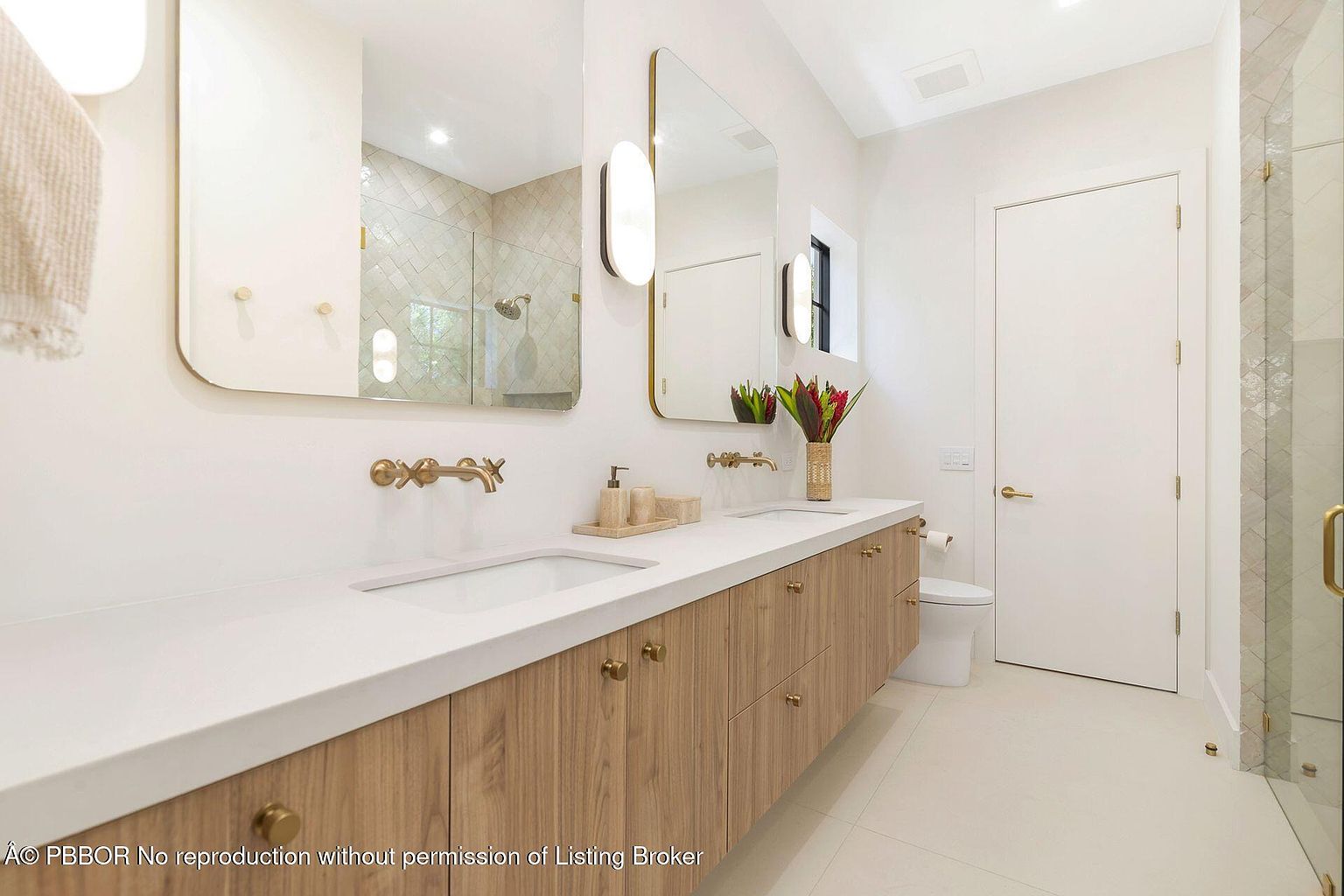 This is a bright and modern primary bathroom featuring a double vanity with a white countertop and light wood cabinetry. The space is illuminated by stylish sconces flanking the mirrors, and gold fixtures add a touch of elegance. The bathroom also includes a toilet and a glass-enclosed shower, creating a clean and luxurious atmosphere.