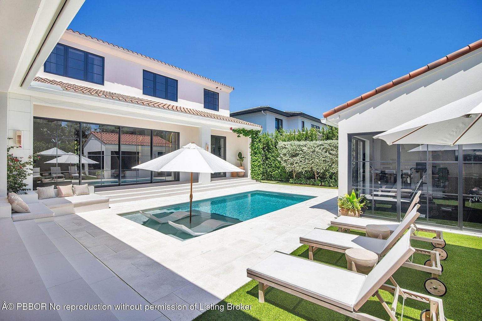 This image showcases a luxurious backyard pool area, featuring a modern rectangular pool with built-in lounge chairs. The surrounding patio is paved with light-colored stone, complemented by elegant lounge furniture and umbrellas. The architecture includes a two-story white house with dark-framed windows and a red tile roof, creating a sophisticated and inviting outdoor living space.
