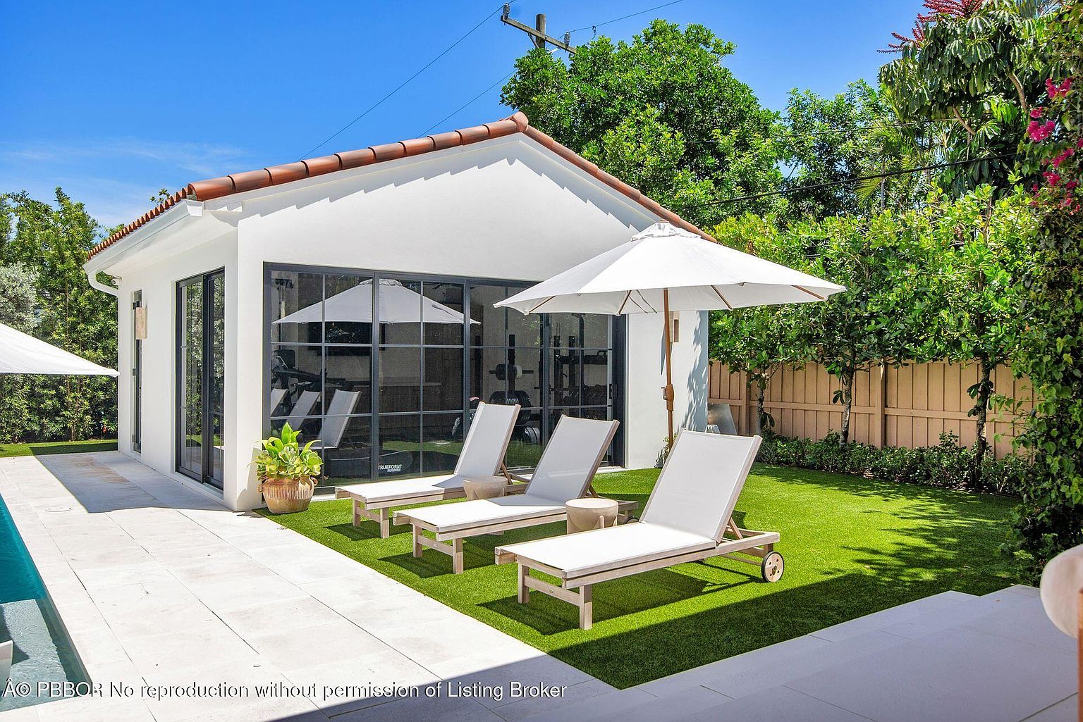 This image showcases a beautifully landscaped backyard featuring a modern pool house with black-framed windows and doors. Three white lounge chairs are arranged on the artificial turf, complemented by a white umbrella for shade. The scene exudes a luxurious and relaxing atmosphere, perfect for outdoor enjoyment.