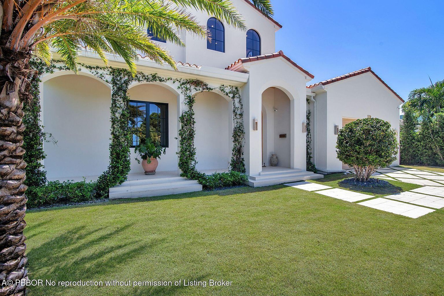 This is a front view of a Mediterranean-style home with a well-manicured lawn. The house features white stucco walls, arched entryways adorned with climbing vines, and a red tile roof. A palm tree is visible on the left, adding to the tropical ambiance.