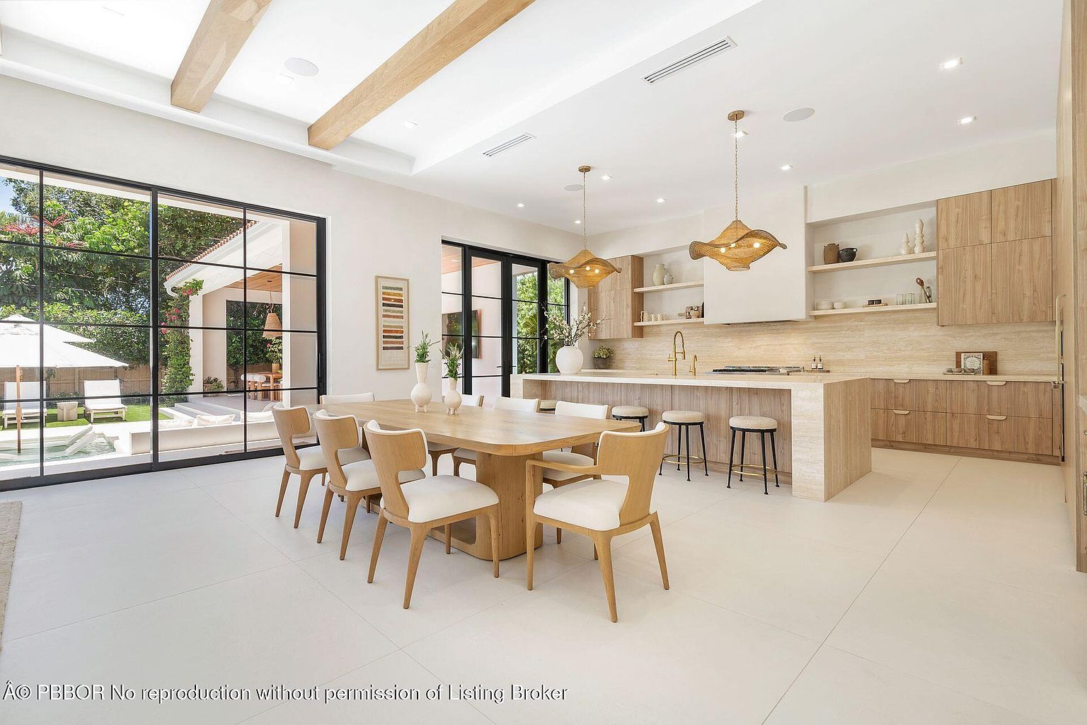 This interior shot showcases a modern kitchen and dining area with a seamless flow. The kitchen features light wood cabinetry, a large island with bar seating, and stylish pendant lighting. The dining area includes a large wooden table surrounded by elegant chairs, with a large window offering a view of the outdoor pool area, creating a bright and airy atmosphere.