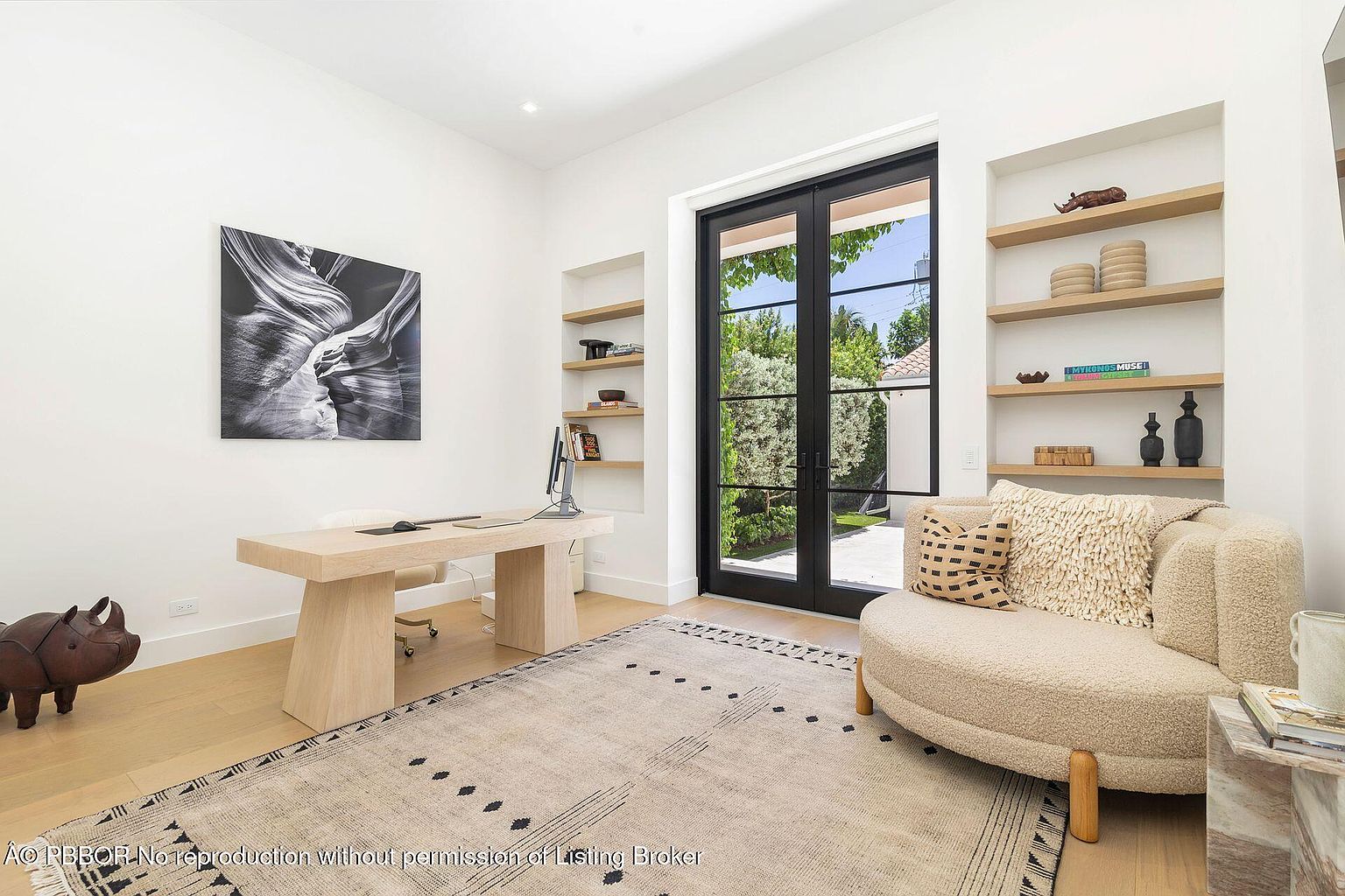 This is an interior shot of a modern office space featuring a light wood desk with a computer, a comfortable round chair, and built-in shelves. A large black-framed glass door provides natural light and a view to the outside. The room is decorated with neutral tones and a patterned rug, creating a serene and stylish workspace.