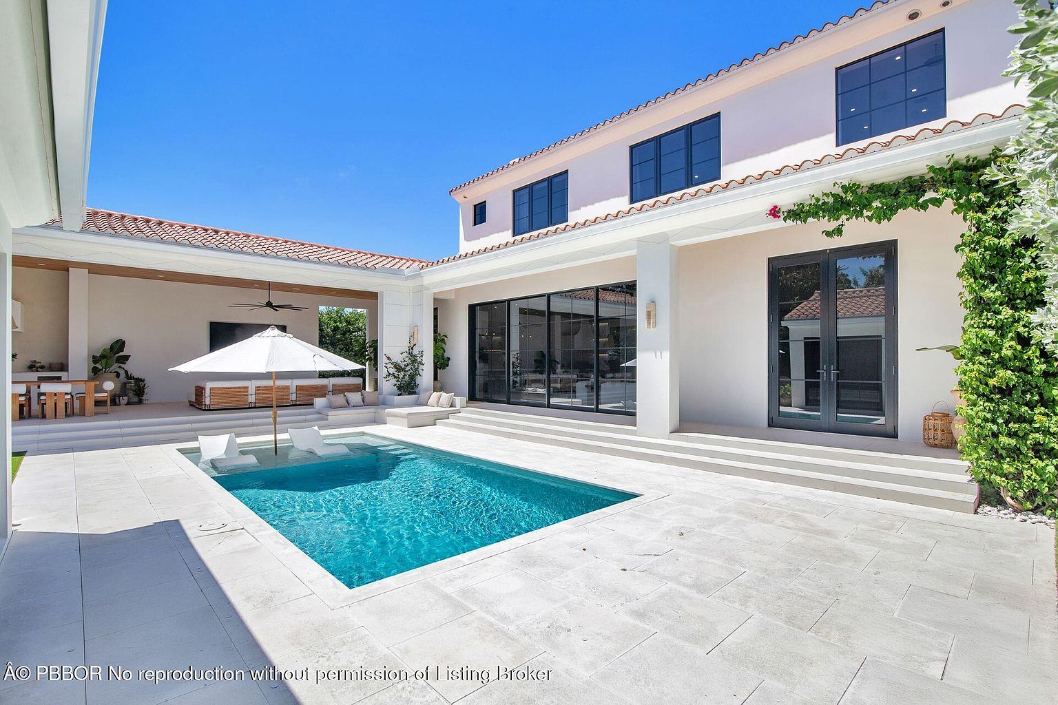 This image showcases a luxurious backyard featuring a rectangular pool with built-in lounge chairs. The pool is surrounded by a light-colored stone patio, and the house has large sliding glass doors that open to the outdoor space. The architecture is modern with clean lines and a light color palette, creating an inviting and upscale atmosphere.