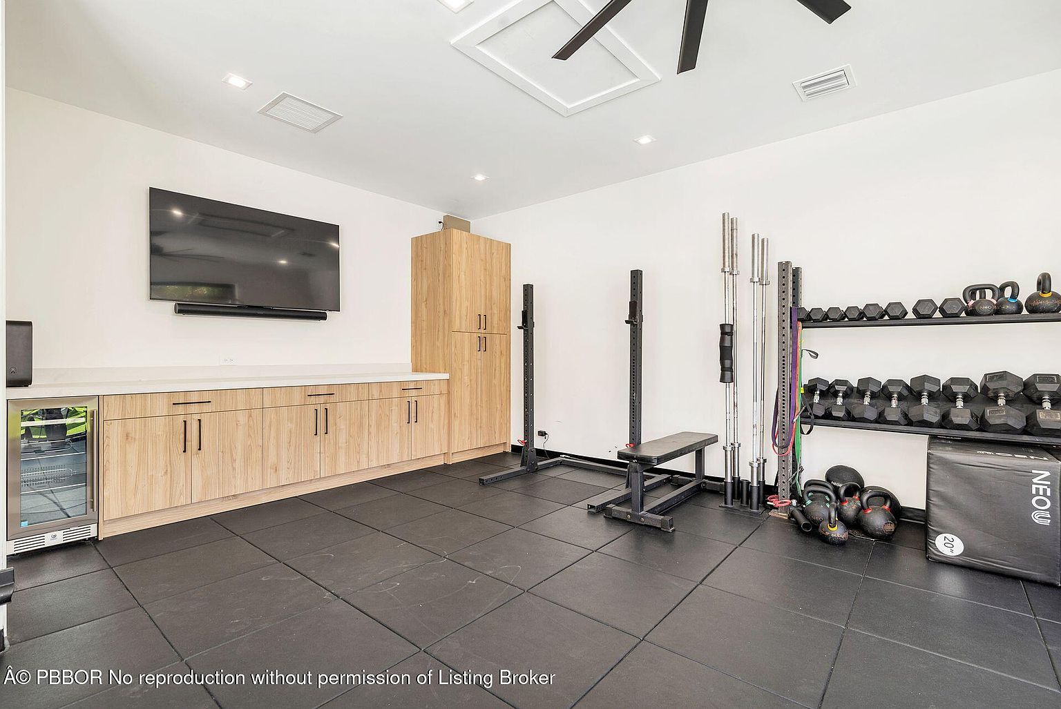 This is an interior shot of a home gym. The room features black rubber flooring, white walls, and a variety of exercise equipment including a weight rack with dumbbells and kettlebells, a squat rack, a bench, and a plyometric box. There is also a built-in cabinet with a beverage cooler and a large television mounted on the wall, suggesting a focus on both fitness and entertainment.